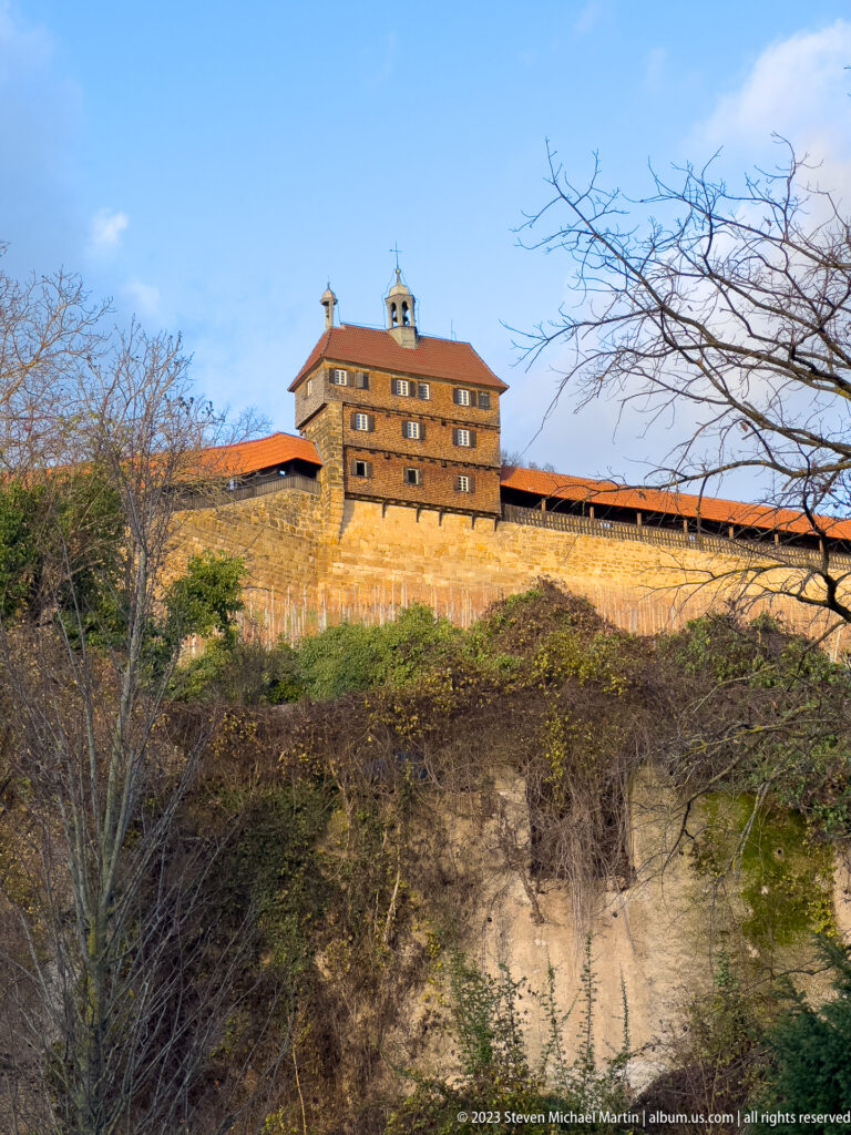Square tower and long covered wall of Esslinger Burg above a steep rough hillside framed by bare branches.