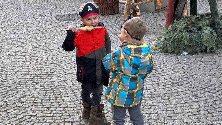 Child in red-and-black pirate costume eating while another child in a plaid jacket stands on cobblestones.