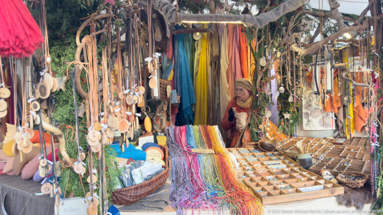 Costumed vendor surrounded by hanging textiles, cords, wooden spoons and small handmade goods in a crowded market stall.