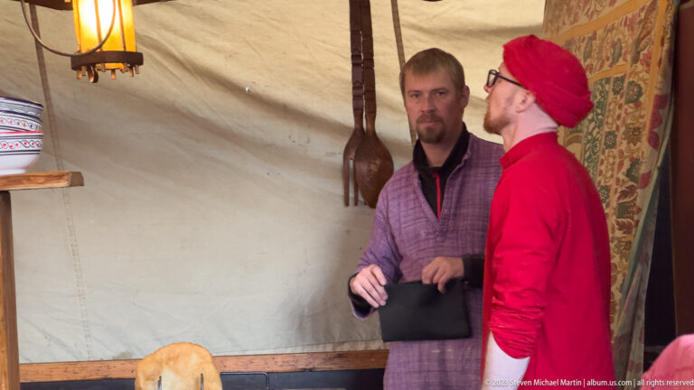 Two men talking inside a canvas market tent beside a hanging long-necked string instrument and patterned textile.