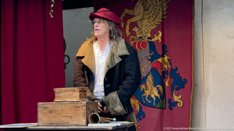 Costumed man in red hat behind wooden boxes and a metal vessel, with a heraldic banner and red curtain behind him.