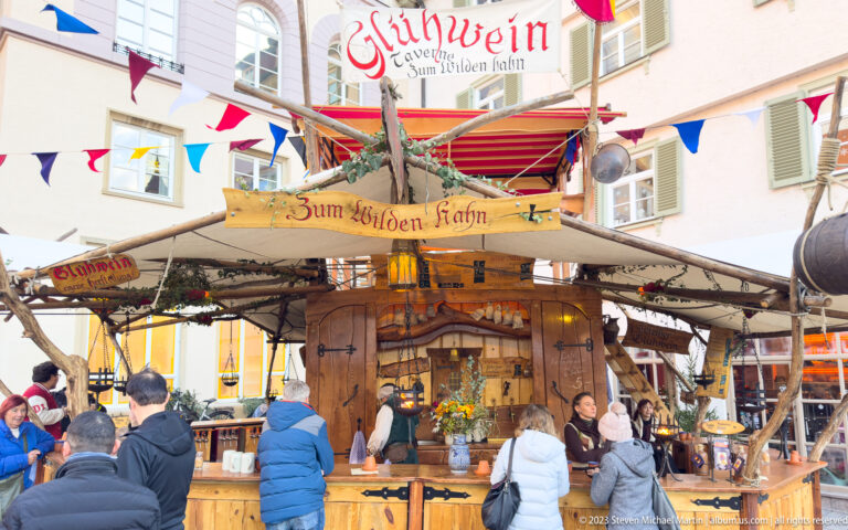 Crowded wooden Glühwein stall marked Zum Wilden Hahn, with bunting and customers in winter clothing.