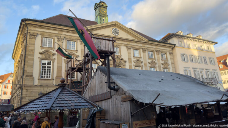 Wooden Christmas market stalls and visitors stand before an ornate pale stone civic building with flags and a clocked pediment.