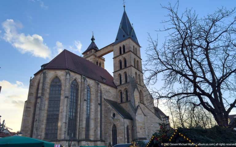 Stone exterior of Stadtkirche St. Dionys with tall Gothic windows, square tower, connecting bridge, bare tree, and Christmas decorations.