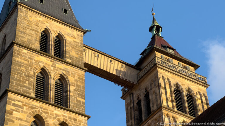 Close upward view of the twin stone towers of Stadtkirche St. Dionys joined by an elevated enclosed bridge.