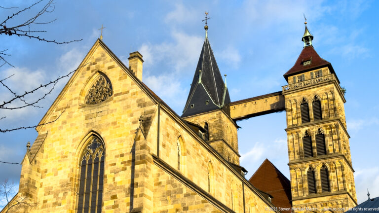 Sunlit stone gable and pointed windows of Stadtkirche St. Dionys with twin towers and their elevated connecting bridge.
