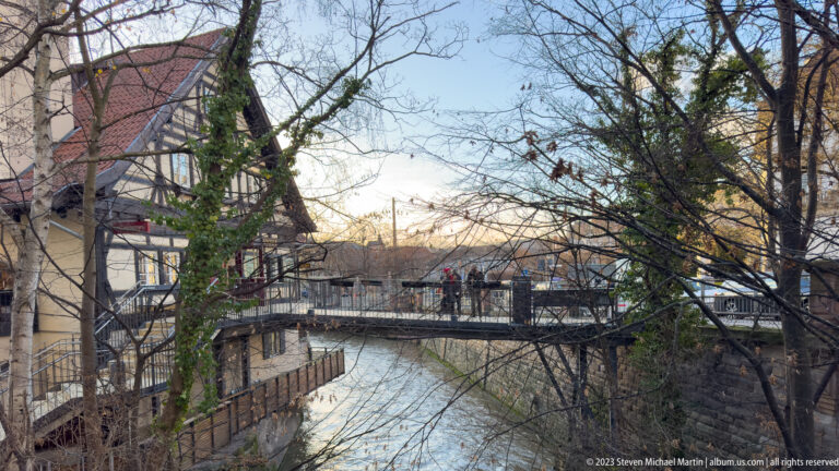 Pedestrians cross a metal bridge over a narrow canal lined by bare trees, stone walls, and historic buildings.