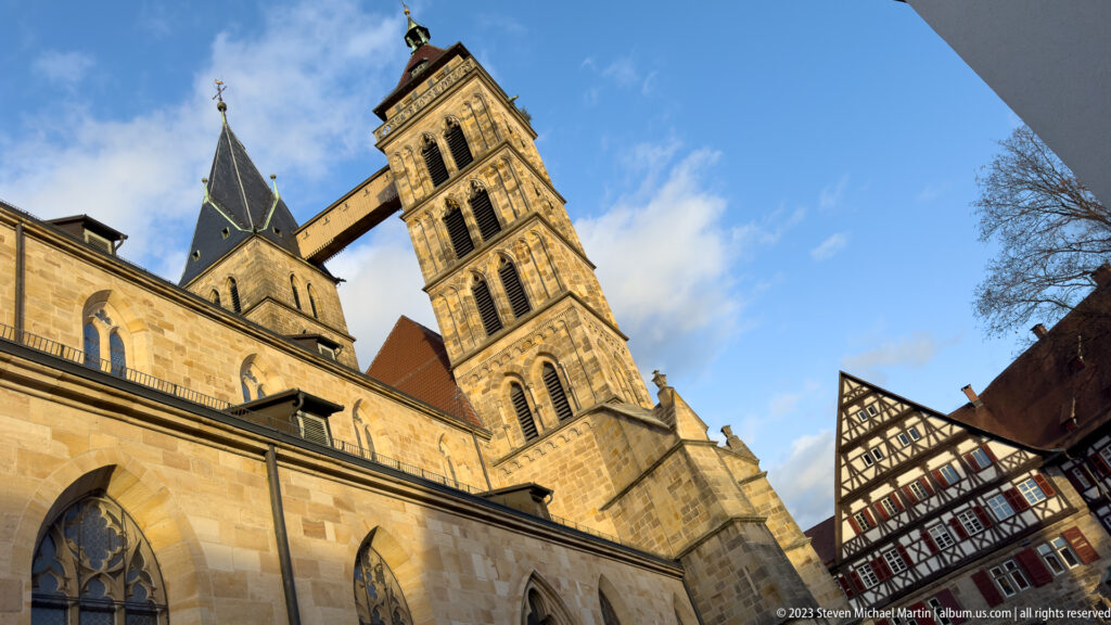 Low-angle view of Stadtkirche St. Dionys showing stone nave, square tower, dark roof, elevated bridge, and adjacent half-timbered house.