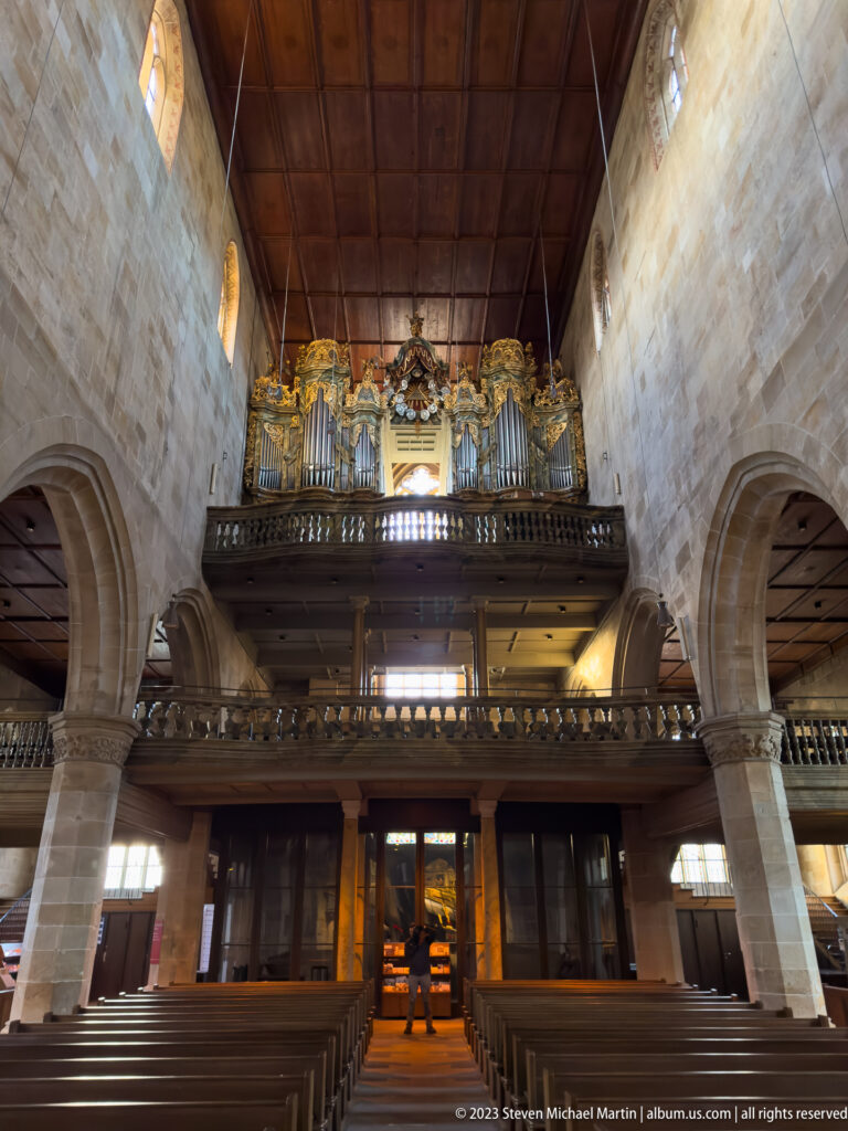 Large pipe organ above a wooden gallery inside a stone church, with pews, arcades, and a broad timber ceiling.