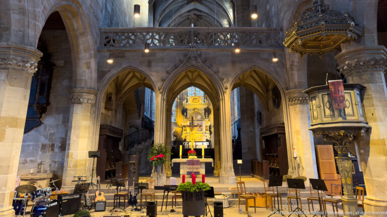 Gothic church chancel with illuminated altar, Advent decorations, carved pulpit, chairs, and musical equipment in the foreground.