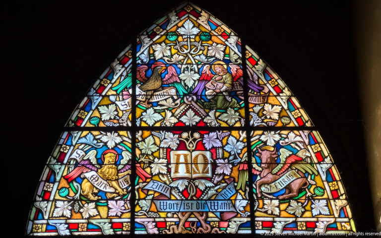 Pointed stained-glass window with colorful figures, foliage, ribbon inscriptions, and a central Christogram against a dark church interior.