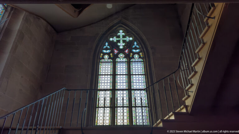 Pointed Gothic window with pale patterned glass beside metal railings and an interior staircase in a stone church.