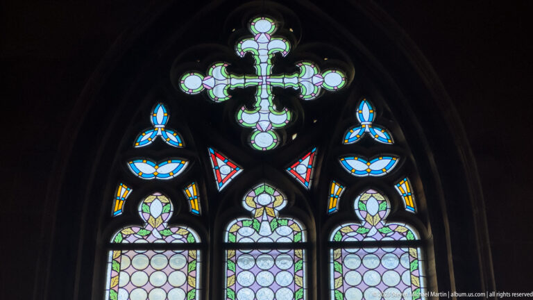 Close view of Gothic stone tracery filled with blue, green, yellow, and pale stained glass around a cross-shaped upper motif.