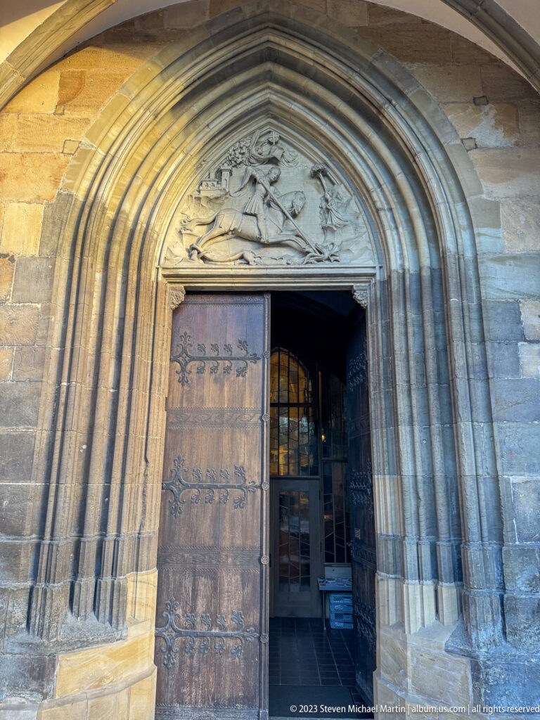 Pointed stone church portal with deep molded jambs, carved tympanum, and a partly open heavy wooden door.