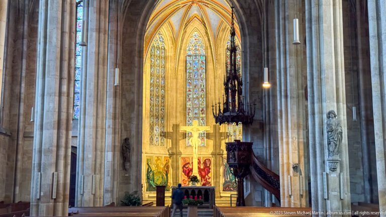 Illuminated Gothic chancel with tall stained-glass windows, central crucifix, colorful altar textiles, and dark carved pulpit.