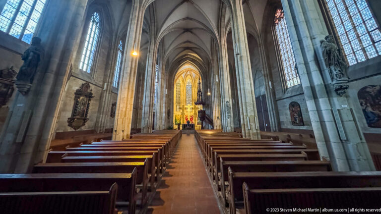 Central aisle through a tall Gothic nave with wooden pews, ribbed vaults, side windows, and a brightly lit chancel in the distance.