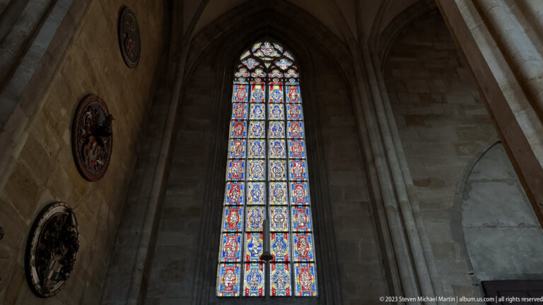 Tall colorful stained-glass window set deep in a pointed stone bay, with circular wall artworks visible at left.