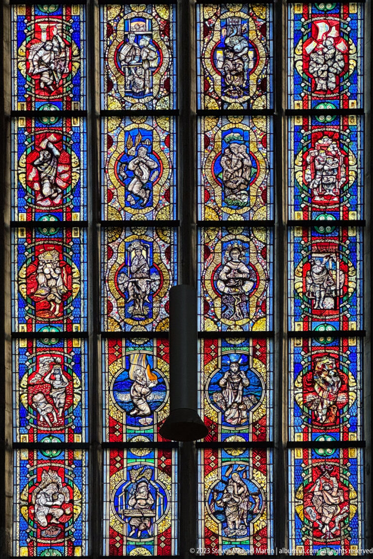 Colorful stained-glass window arranged in vertical narrative panels, partly obscured by a dark hanging light fixture.