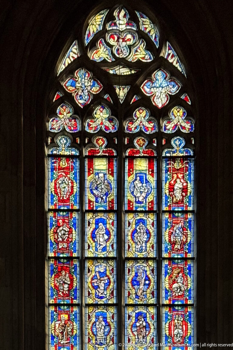 Large Gothic stained-glass window with rows of colorful figurative panels and ornate floral tracery glowing in a dark interior.