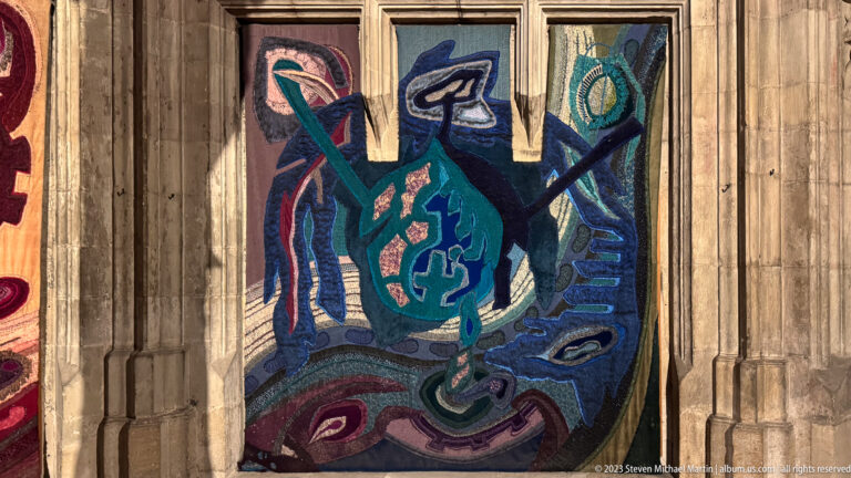 Abstract blue-green woven textile panel hanging between slender Gothic stone shafts inside a church.