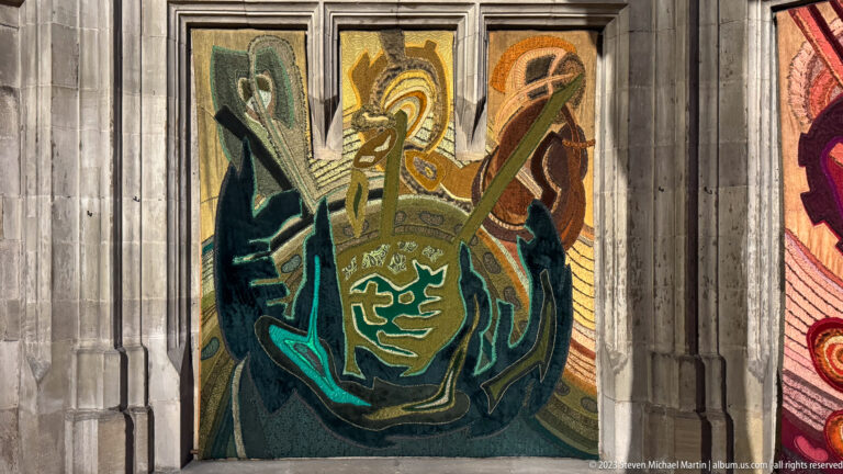 Green, turquoise, ochre, and brown abstract woven textile displayed within a narrow Gothic stone bay.