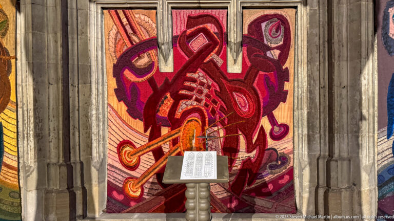 Red and magenta abstract woven textile behind a lectern with an open book, framed by pale Gothic stone supports.