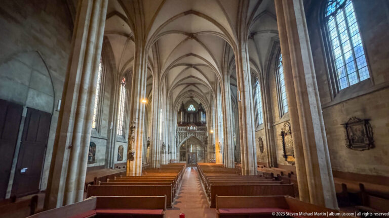 Long Gothic church nave with central aisle, wooden pews, ribbed vaults, tall columns, side windows, and distant organ gallery.
