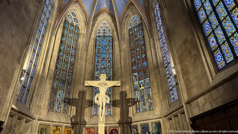 Large crucifix centered before tall colorful stained-glass windows in a high Gothic church chancel.