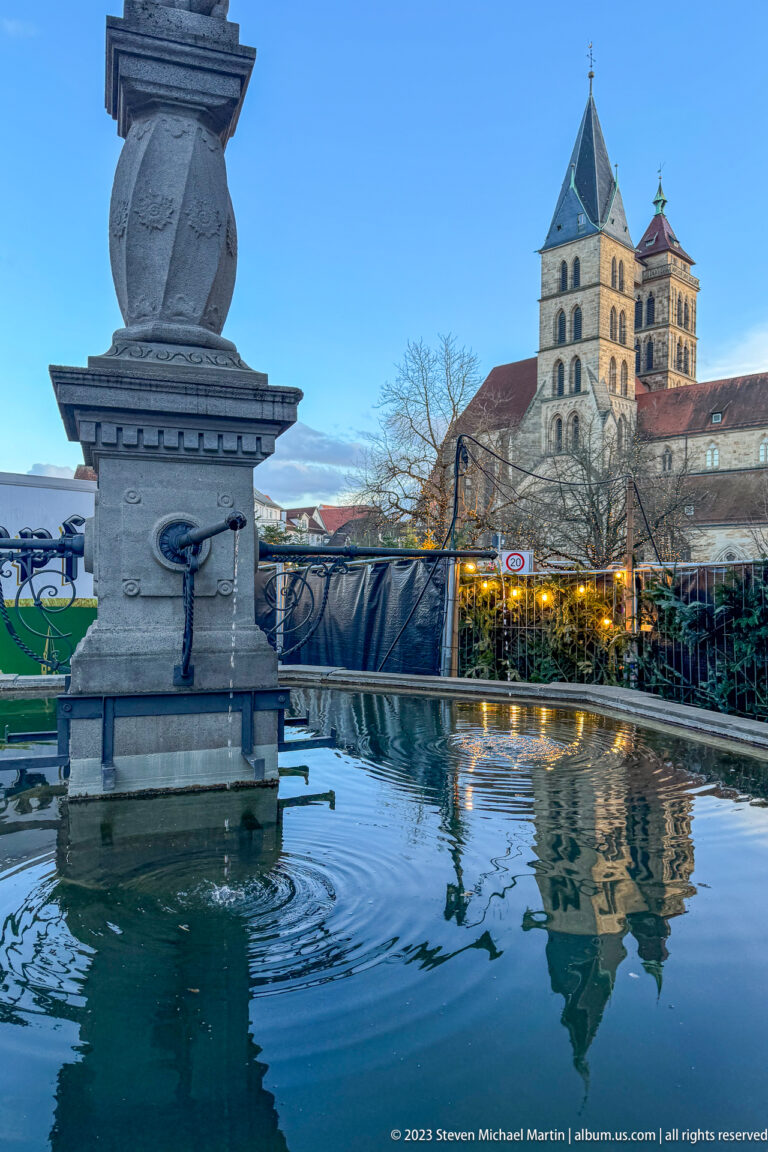 Stone fountain basin reflecting Stadtkirche St. Dionys, with winter market lights and bare trees beyond.
