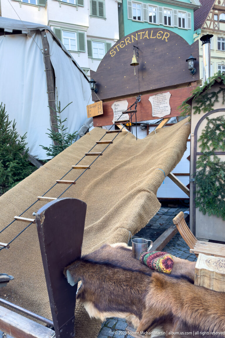 Large sloping fabric-covered wooden market prop beneath a Sternentaler sign, with hides and evergreen branches nearby.