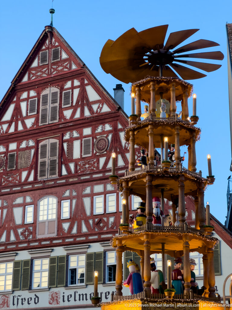 Large illuminated tiered Christmas pyramid with rotor blades in front of a red-and-white half-timbered house at dusk.