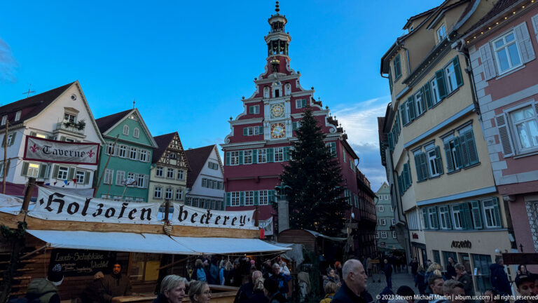 Red stepped-gable Altes Rathaus with clock faces above a crowded Christmas market and timber bakery stall.