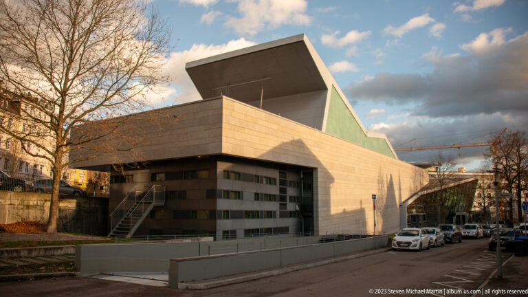 Angular pale-stone modern public building with projecting upper volume, exterior stairs, parked cars, and bare trees.