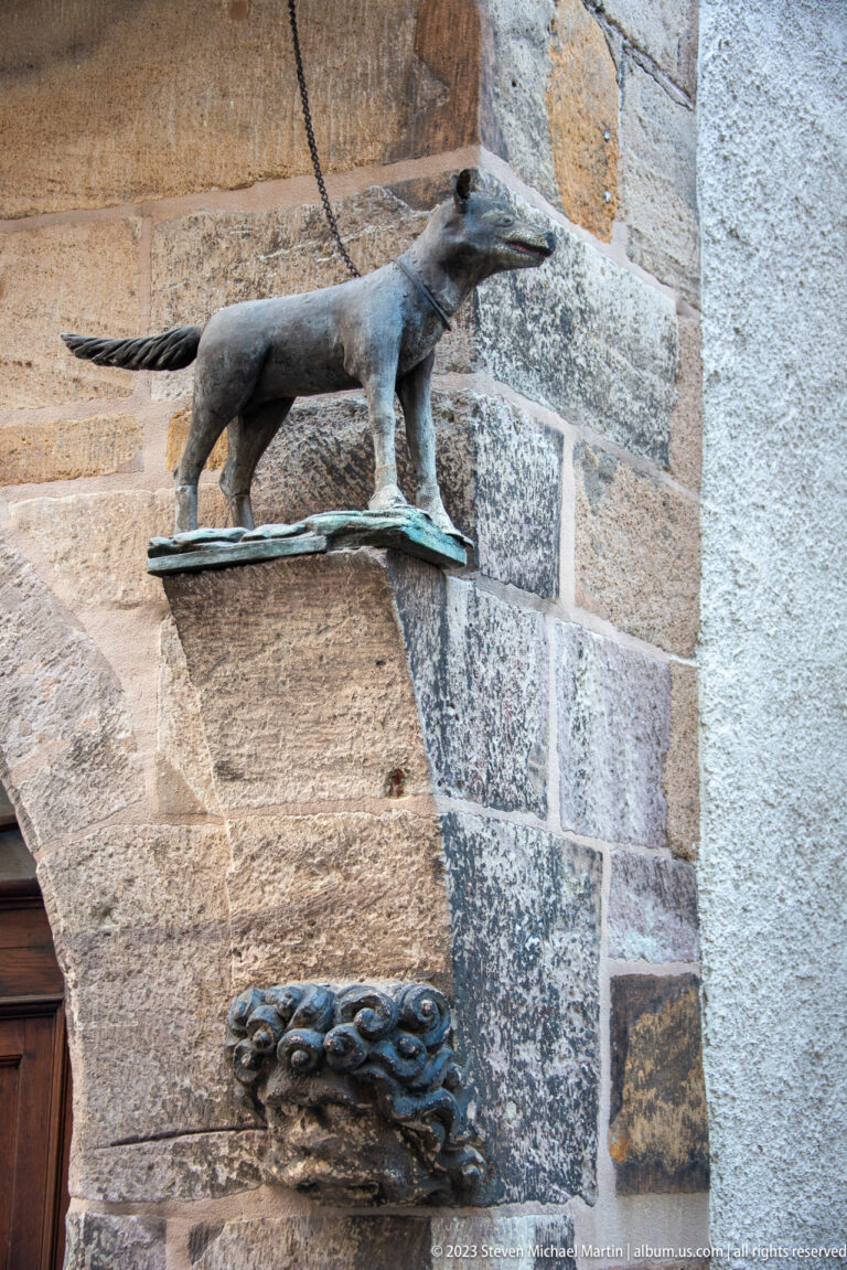 Small bronze animal figure with long tail mounted on a stone wall above a separate flowing metal ornament.
