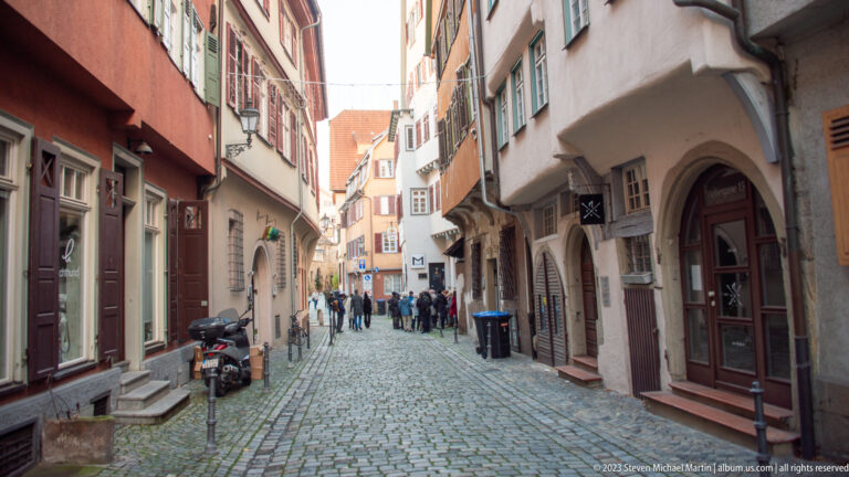Narrow cobblestone street lined with plastered historic houses, shutters, projecting upper stories, and pedestrians in the distance.
