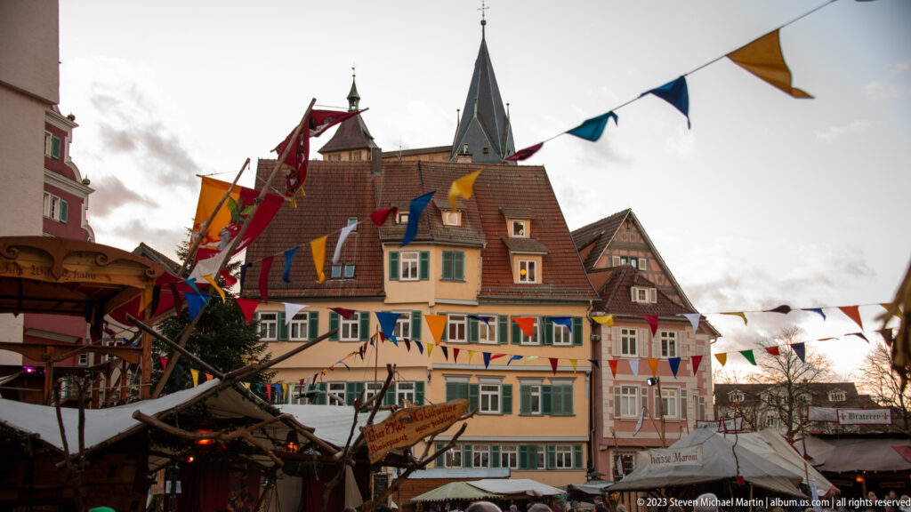 Christmas market stalls and colorful pennants beneath historic houses, with a slender church spire rising behind the tiled roofs.