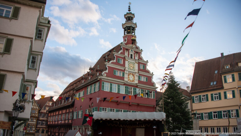 Oblique low-angle view of the red Altes Rathaus with stepped gable, clock, green shutters, lantern, and market pennants.