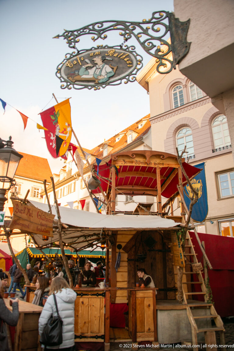 Rustic two-level Christmas market booth with exterior stairs, red-and-gold canopy, flags, and wrought-iron overhead sign.