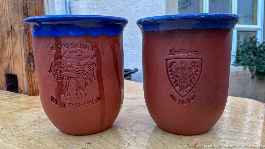 Two reddish ceramic Christmas market cups with blue rims, including one embossed with the Esslingen am Neckar crest.