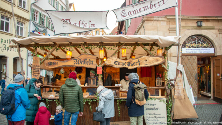 Wooden Christmas market food stall with hand-painted waffle and Camembert signs, warm lamps, greenery, and waiting customers.