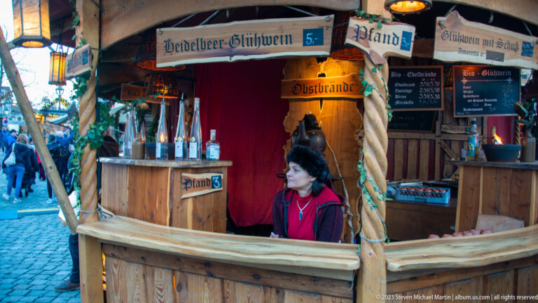 Vendor inside a rustic wooden Glühwein booth with menu boards, bottles, garlands, and hand-painted signs.