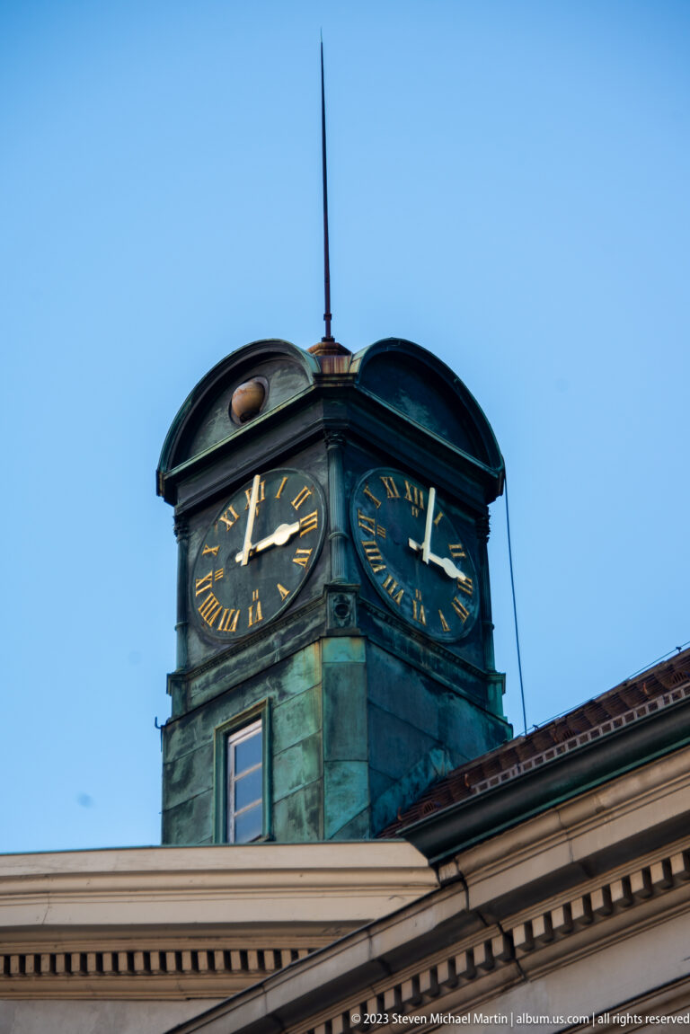 Green-patinated rooftop clock turret with gilded Roman numerals and hands above a pale classical cornice.