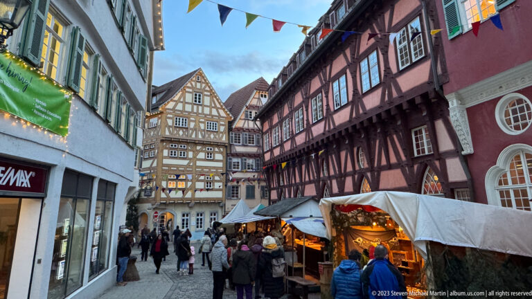 Pedestrian Christmas market lane with canvas-covered stalls beside dark-red half-timbered buildings and a patterned gabled house ahead.