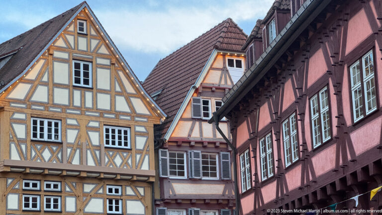 Cluster of steep-gabled half-timbered houses with contrasting ochre, pale, and dark-red timber patterns and small-paned windows.