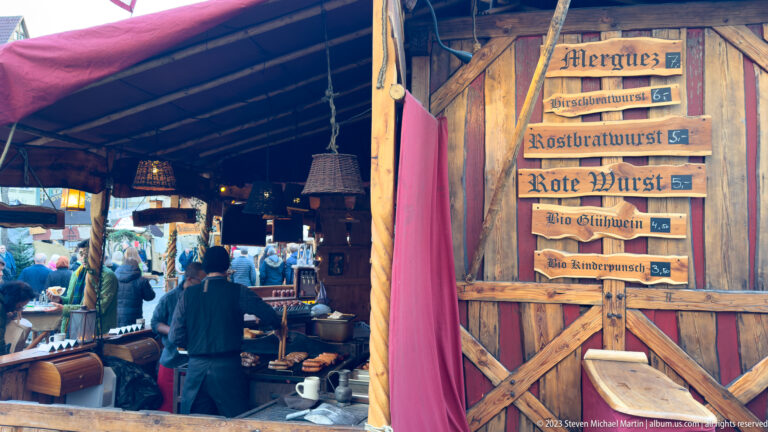 Interior of a rustic wooden food stall with customers and a painted German sausage menu on the door.