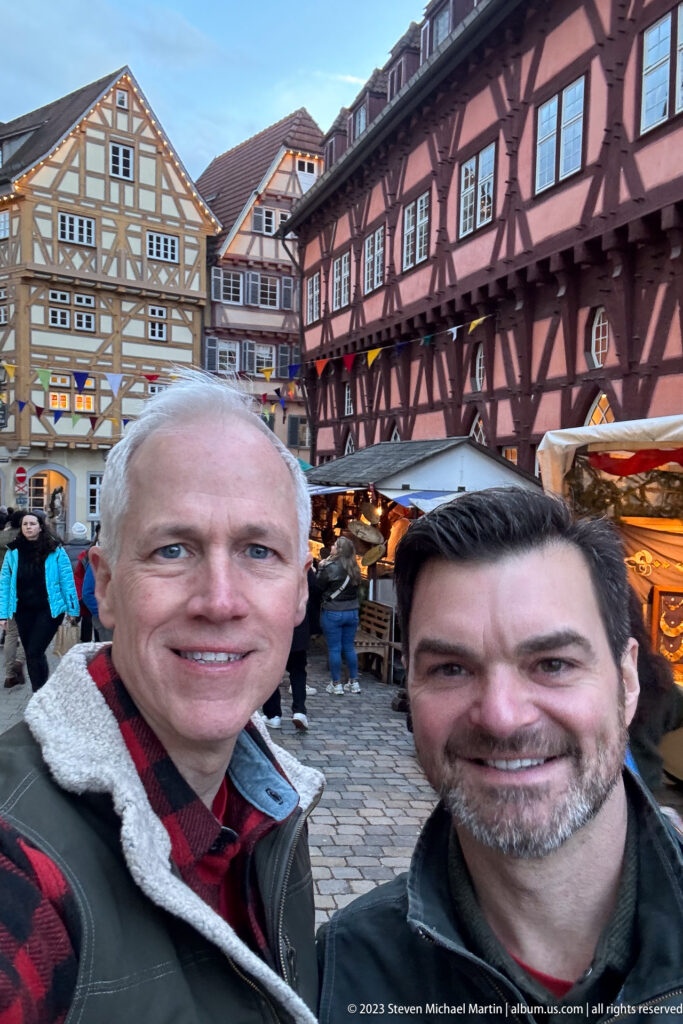Two smiling adult men taking a selfie in a Christmas market with half-timbered buildings and stalls behind them.