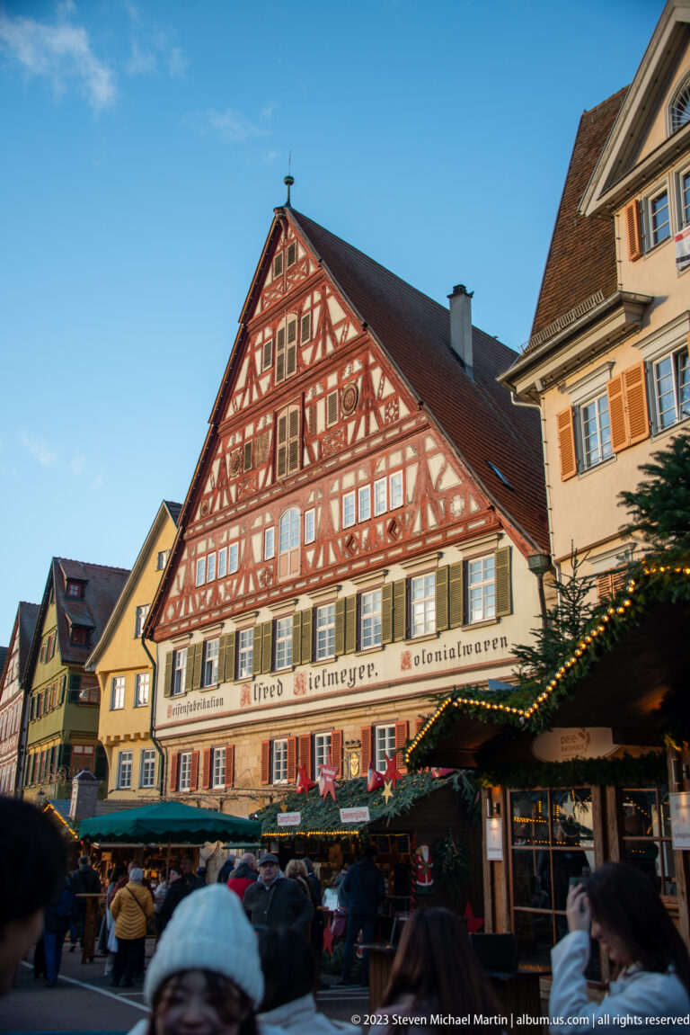 Large red-brown half-timbered gabled building above Christmas market stalls, greenery, and visitors in warm late-day light.