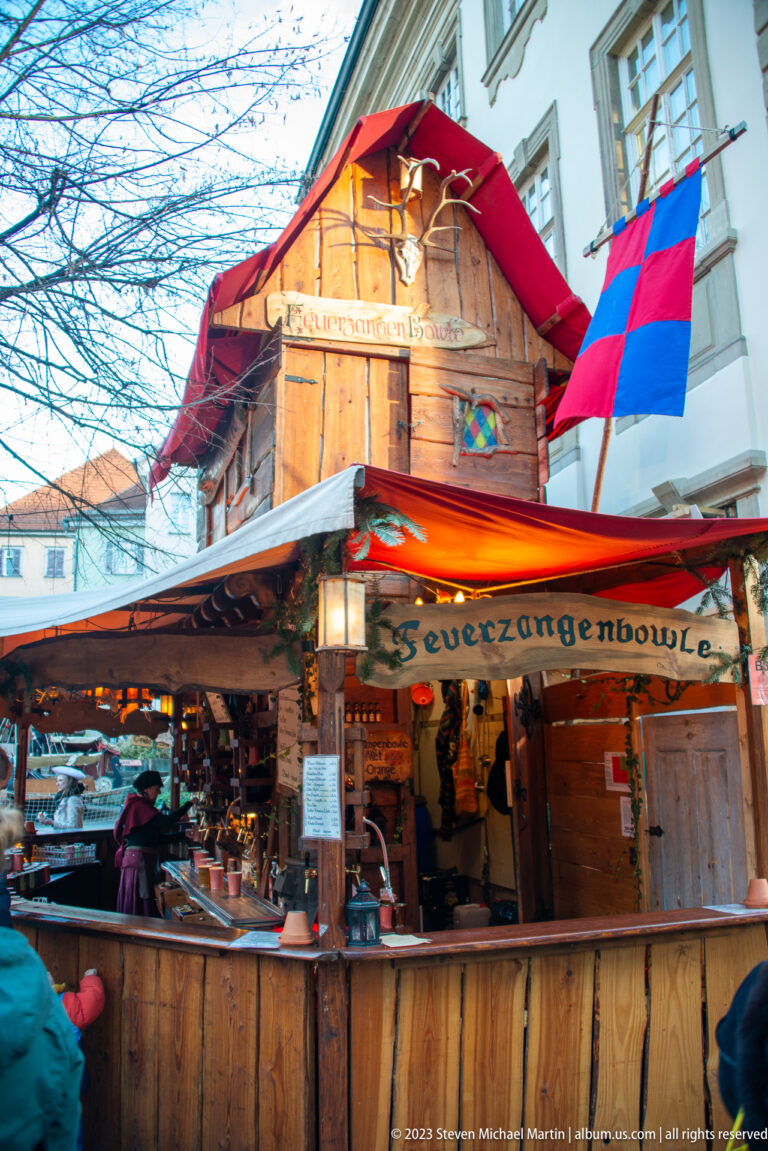 Wooden Feuerzangenbowle Christmas market booth with red-and-blue flags, warm lamps, bottles, and visitors at the counter.