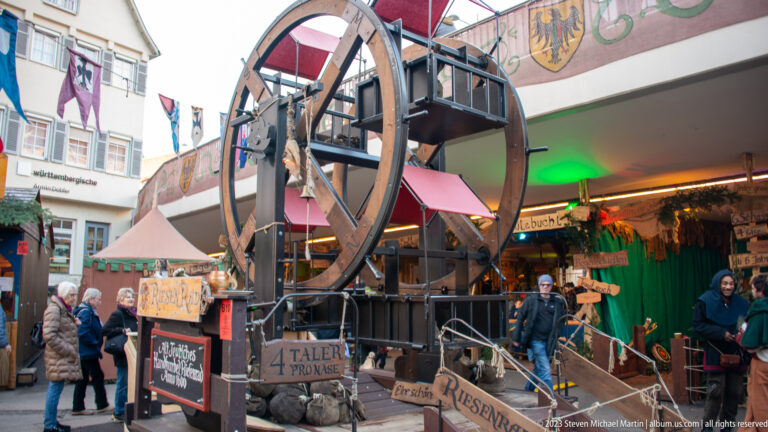 Large vertical wooden wheel beside stairs and timber platforms at a medieval-themed Christmas market stall with visitors.