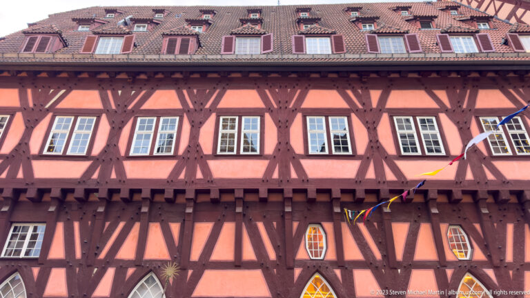Close view of salmon-pink half-timbered façade with dark braces, white windows, colored panes, dormers, and red tiled roof.